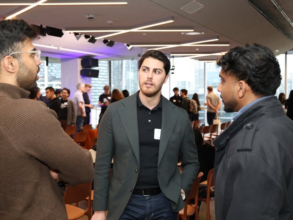 A group of people are gathered in a modern indoor space for a networking event, with three men in the foreground engaged in a conversation.
