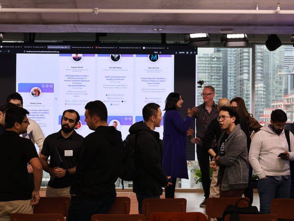 A group of people are gathered in a modern indoor space for a networking or demo event, with a large screen in the background displaying project profiles.