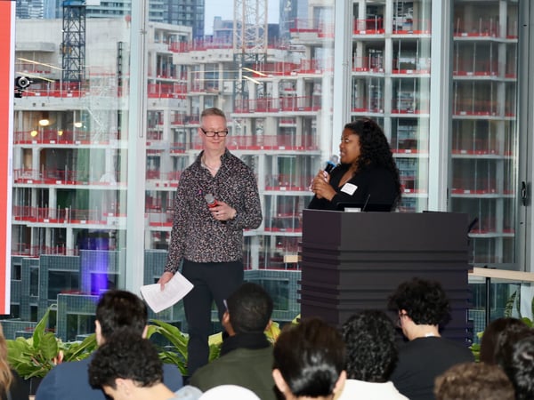 A woman speaks at a podium with a microphone while a man stands beside her holding papers, in front of an audience and a large window overlooking a city under construction.