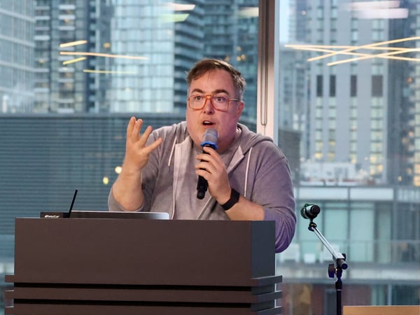A man in a grey hoodie and orange-rimmed glasses is speaking into a microphone while standing behind a podium, with a city skyline visible through a large window in the background.
