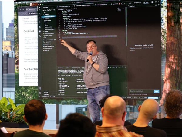 A man stands in front of a large screen displaying code, delivering a presentation to an audience in a modern indoor space with city views.