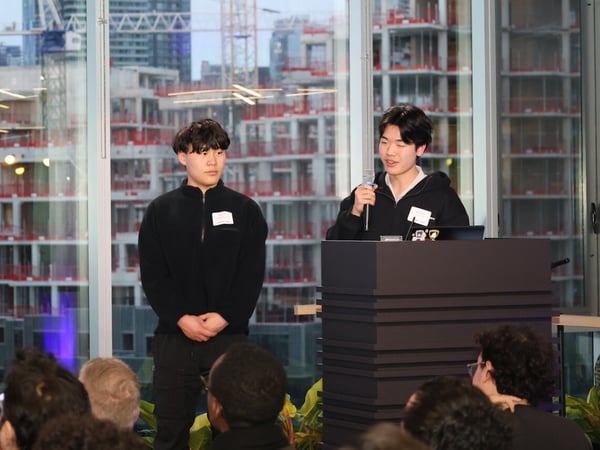 Two young men are giving a presentation at a podium in front of an audience, with a large window showing a city construction site in the background.