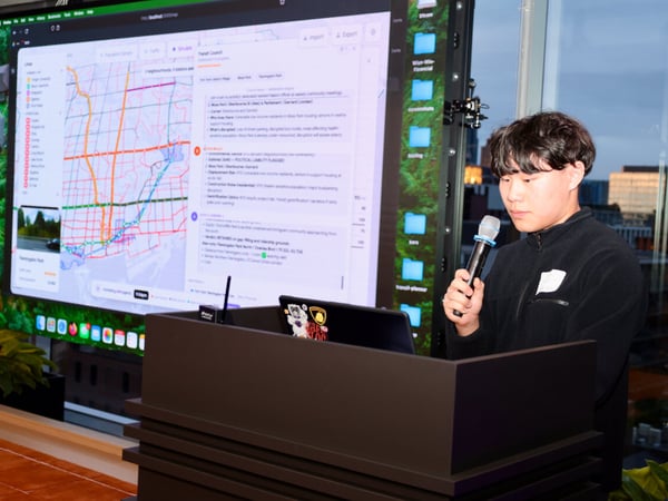 A young man is giving a presentation at a podium, holding a microphone, with a large screen behind him displaying a map-based transit planning interface.