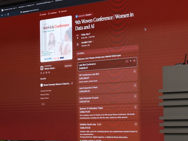 A photograph of a large digital display screen showing a ticket registration page for the '9th Woven Conference | Women in Data and AI' on the Luma website.