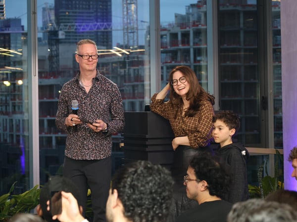 A man and a woman stand in front of a large window with a city skyline view, appearing to host an event or give a presentation to an audience.