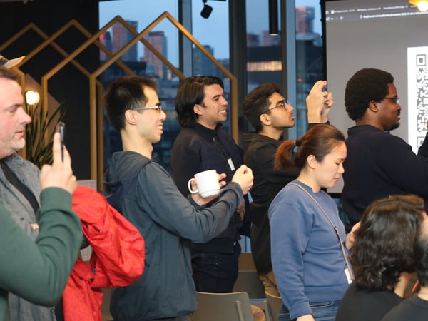 A group of people at an event or office are looking at a screen displaying a large QR code, with several individuals using their smartphones to scan it.
