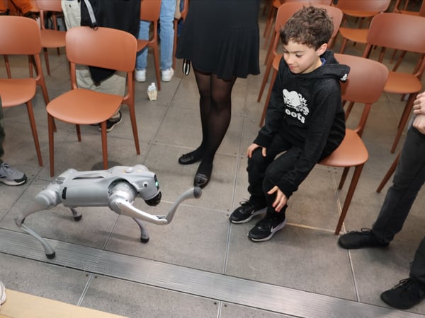 A young boy sitting on a chair watches a silver quadruped robot with an attached robotic arm in a room filled with brown chairs and several standing adults.
