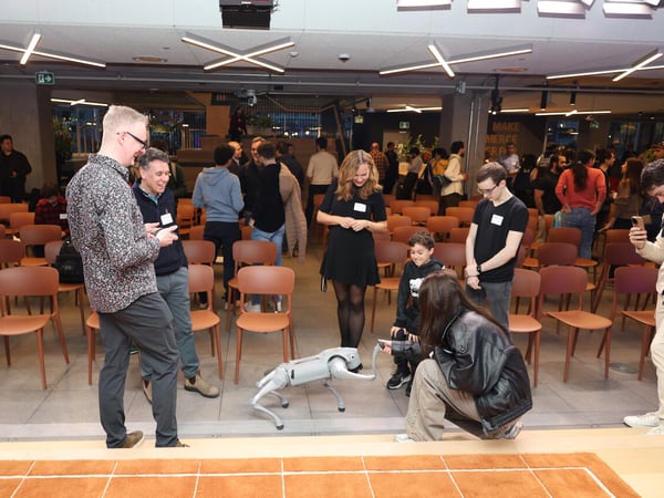 A group of people in a modern office or event space are gathered around to watch a demonstration of a four-legged robotic dog.