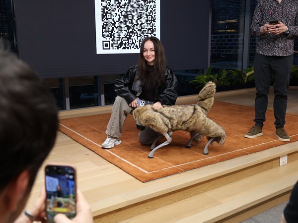 A woman kneels on a brown rug to pet a furry robotic dog during a technology demonstration, with a large QR code displayed on a screen behind her.