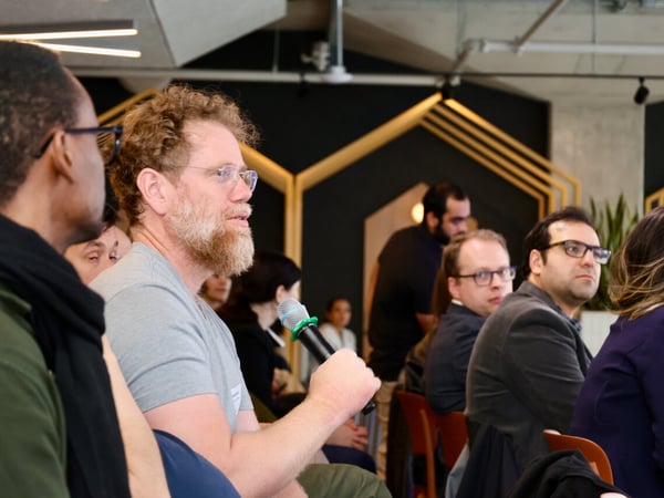 A man with a beard and glasses holds a microphone, speaking during an indoor event or discussion while others listen in the background.