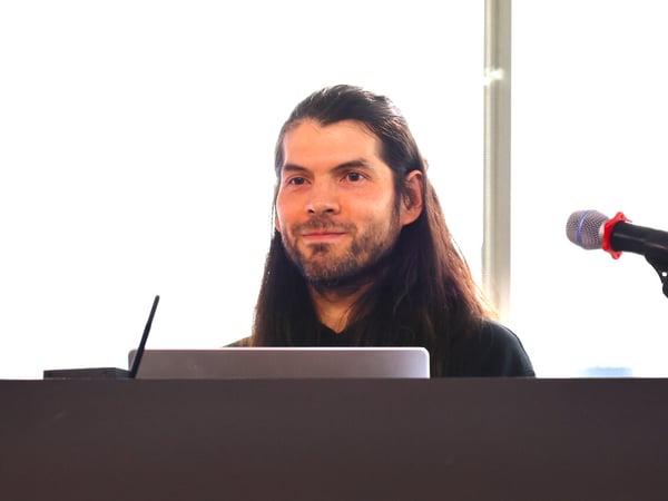 A man with long dark hair and a beard smiles while standing behind a podium with a laptop and a microphone.