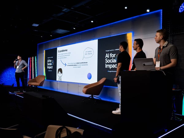 A group of young men are giving a presentation on a stage titled 'AI for Social Impact' during a Google Hackathon.