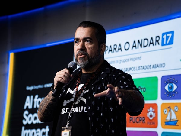 A man with a beard and tattoos is speaking into a microphone while giving a presentation in front of a digital screen.