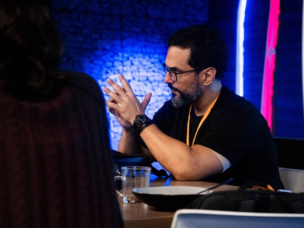 A man with a beard and glasses is captured in profile, gesturing with his hands during a conversation at a table in a dimly lit room with neon accents.