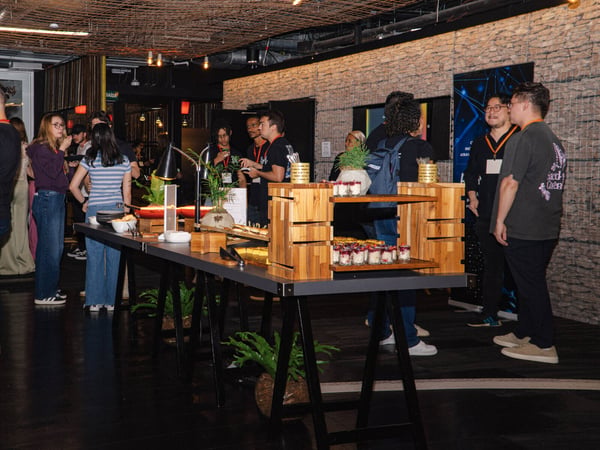 A group of people are gathered around a long table with snacks and desserts at an indoor social event.