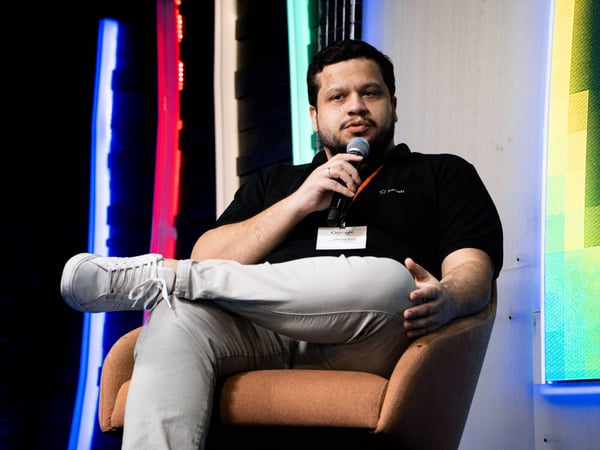 A man is sitting in an orange armchair on a stage, holding a microphone and speaking during an event.