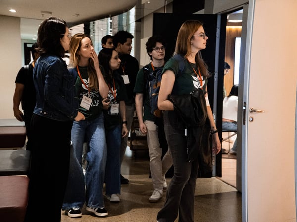 A group of young adults, some wearing matching green t-shirts and orange lanyards, are gathered in an indoor hallway near an open doorway.