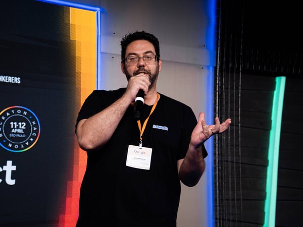 A man with glasses and a beard is speaking into a microphone during a presentation at a hackathon event in São Paulo.