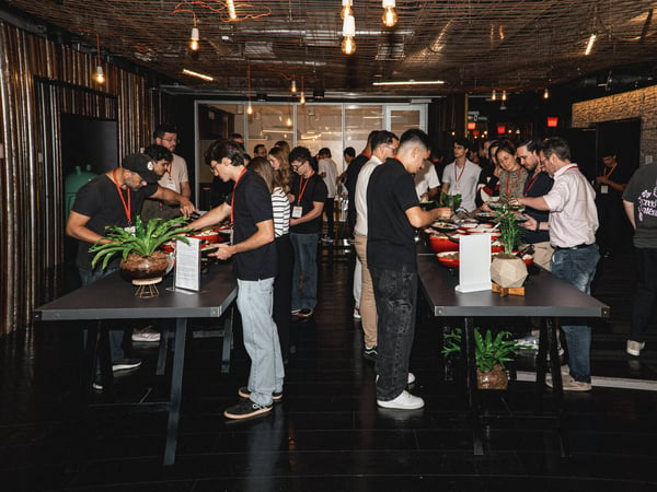 A group of people are gathered in a modern, industrial-style indoor space, serving themselves from a buffet laid out on two long tables.
