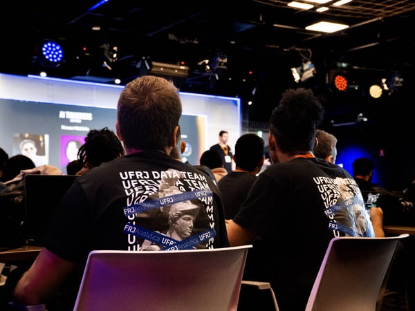 A photograph taken from the back of an auditorium showing an audience watching a presentation, with two people in the foreground wearing 'UFRJ DATA TEAM' t-shirts.