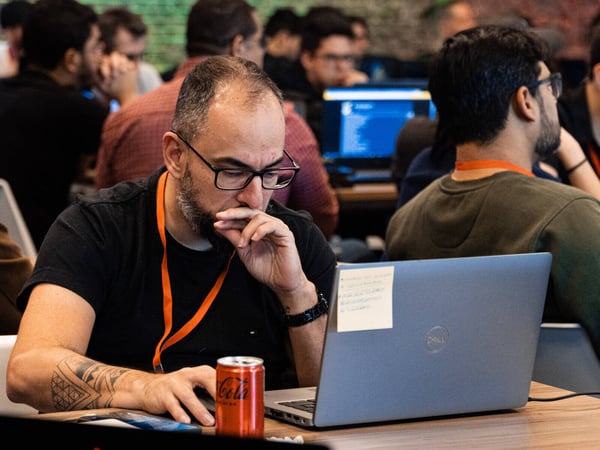 A man with glasses and a beard is deeply focused on his laptop in a busy indoor workspace, surrounded by other people working.