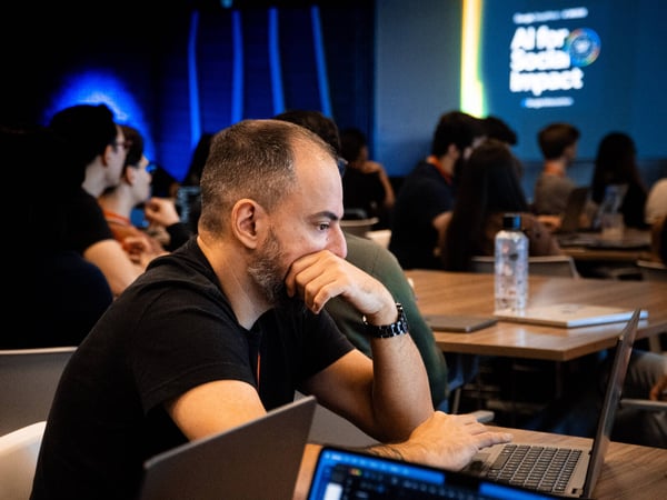 A man in a black t-shirt sits at a wooden table with a laptop, looking thoughtfully towards a presentation screen in a dimly lit room.