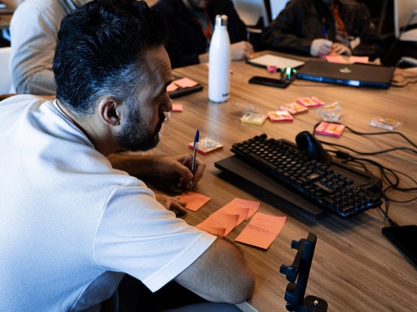 A man in a white shirt is focused on writing on orange sticky notes at a wooden table during a collaborative workshop or brainstorming session.