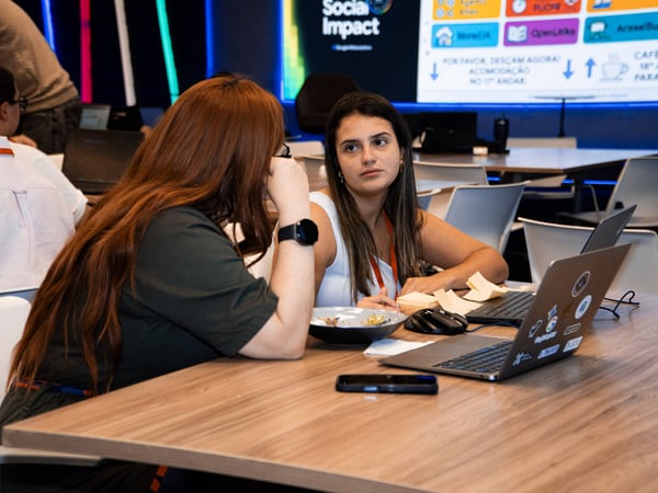 Two women are engaged in a discussion at a wooden table in a modern workspace, with laptops and sticky notes in front of them and a large informational screen in the background.