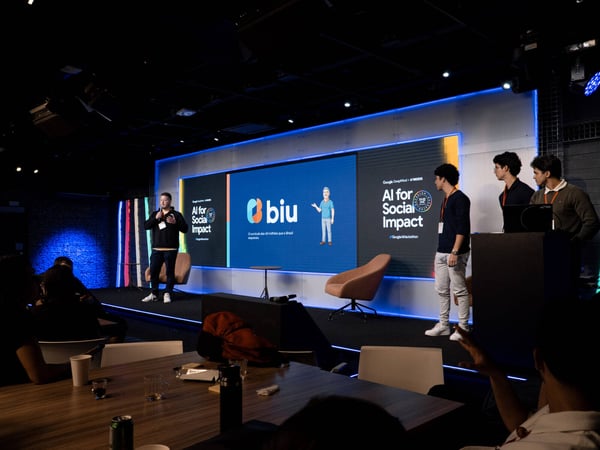 A man is delivering a presentation on a stage during an 'AI for Social Impact' event, with a large digital screen behind him displaying the 'biu' logo and Portuguese text.