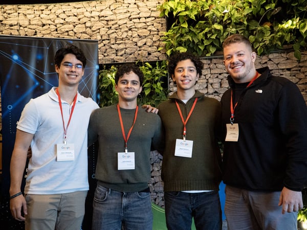 Four young men are standing side-by-side and smiling at a Google event, all wearing identification badges with red lanyards.