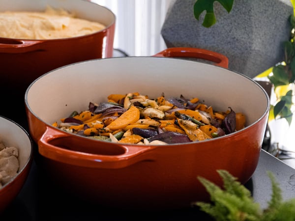 A close-up photograph of a vibrant orange enameled Dutch oven filled with roasted vegetables, including carrots and red onions.