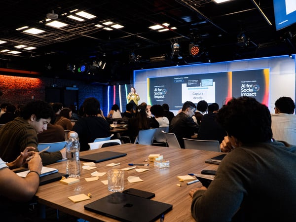 A woman delivers a presentation titled 'AI for Social Impact' to an audience seated at tables in a large, dimly lit event space.