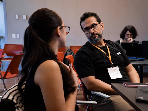 A man wearing glasses and a Google lanyard is engaged in a conversation with a woman in an indoor setting.
