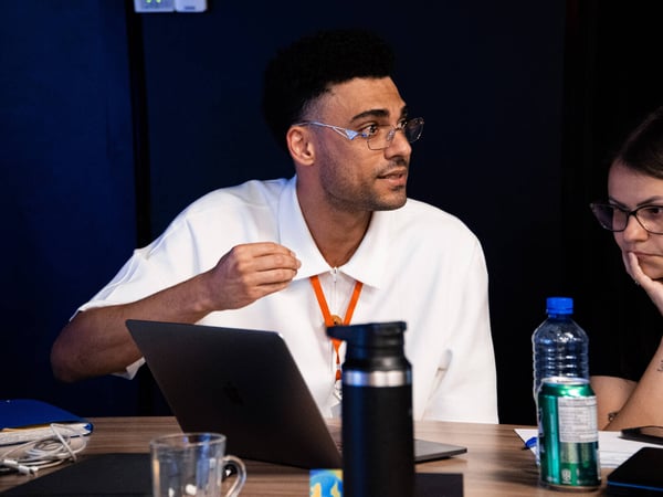 A man in a white polo shirt and glasses gestures while speaking during a meeting or discussion at a table with a laptop and drinks.