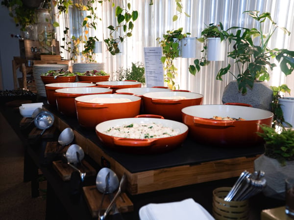 A buffet table setup featuring several large orange ceramic pots filled with food, accompanied by serving spoons and decorative indoor plants.
