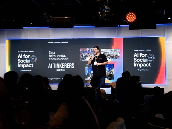 A man stands on a stage giving a presentation in front of a large digital screen displaying 'AI for Social Impact' and 'AI TINKERERS SÃO PAULO' to a silhouetted audience.