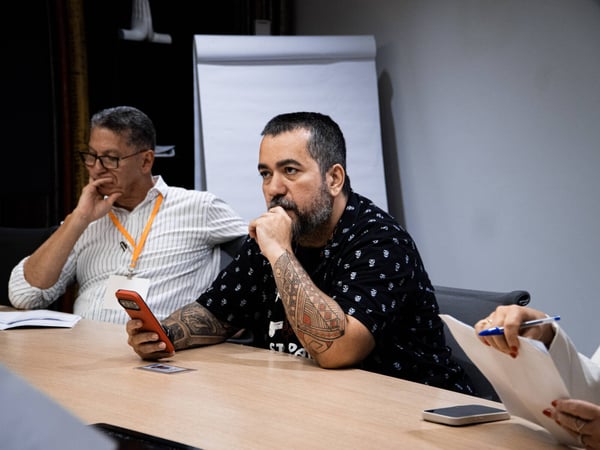 A group of individuals are seated around a wooden table in a meeting or conference setting, appearing engaged and thoughtful.