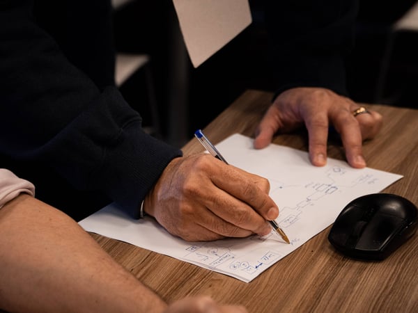 A close-up photo of a person's hands drawing a technical workflow diagram on a piece of paper with a pen on a wooden desk.