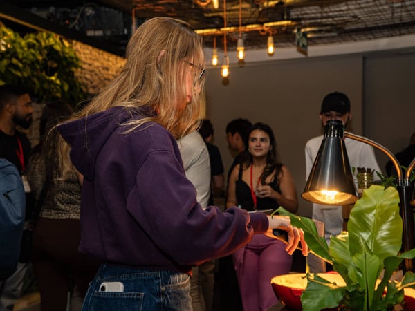 A candid photograph of people at an indoor social event, with a woman in a purple hoodie in the foreground looking at a food table.