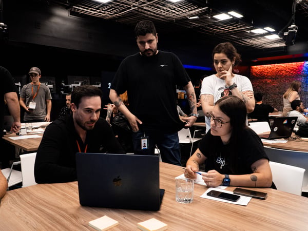 A group of four people in a modern office environment are gathered around a laptop, focused on the screen in a collaborative discussion.