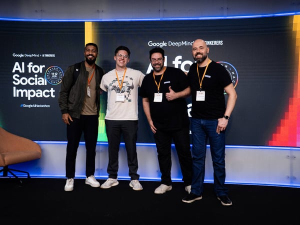 Four men pose for a group photo in front of a large digital screen at a Google AI Hackathon event.