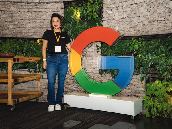 A woman stands posing next to a large, three-dimensional Google 'G' logo in an indoor setting with a stone wall and plants.