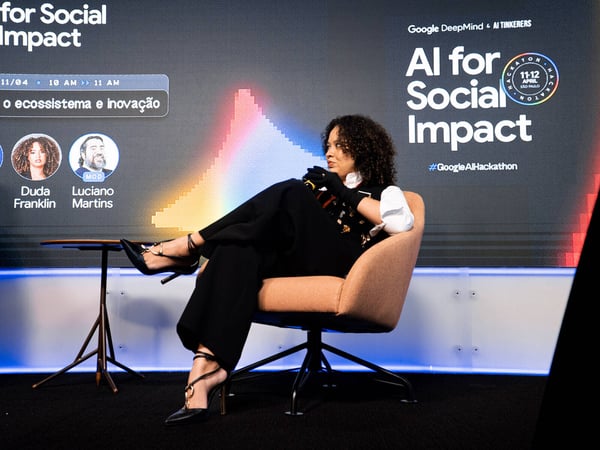 A woman sits on a stage in front of a large digital screen displaying information for an 'AI for Social Impact' event hosted by Google DeepMind and AI Tinkerers.
