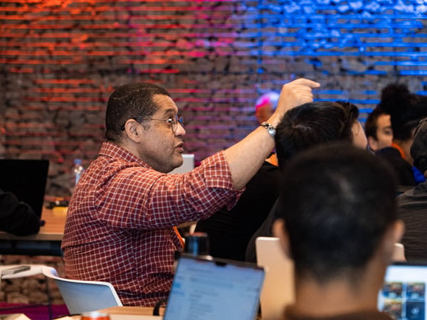 A man in a red plaid shirt raises his hand to ask a question during a workshop or presentation in a room with a brick wall lit by red and blue lights.