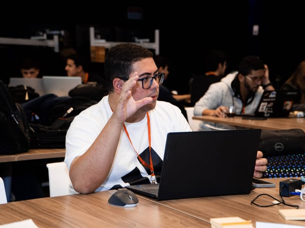 A man in a white t-shirt and glasses is sitting at a wooden desk with a laptop, gesturing with his hand during a conversation in a coworking space.
