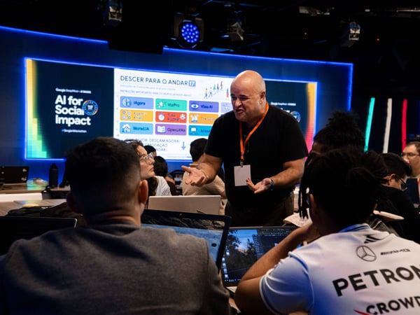A man stands and gestures while speaking to a group of people seated at tables with laptops during an 'AI for Social Impact' event.