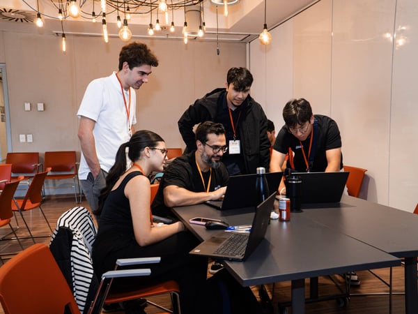 A group of five individuals are gathered around a table with laptops in a modern indoor setting, appearing to be engaged in a collaborative project or workshop.