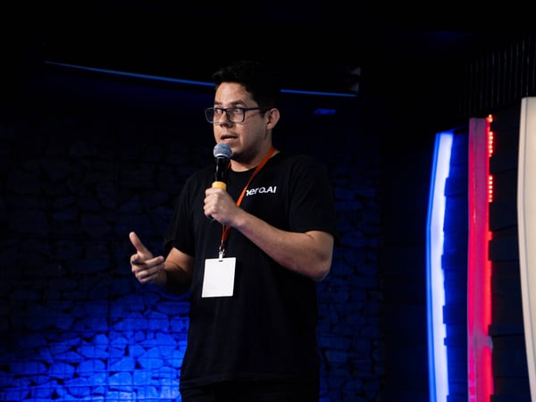 A man wearing glasses and a black t-shirt is speaking into a microphone on a stage with dramatic blue and red lighting.