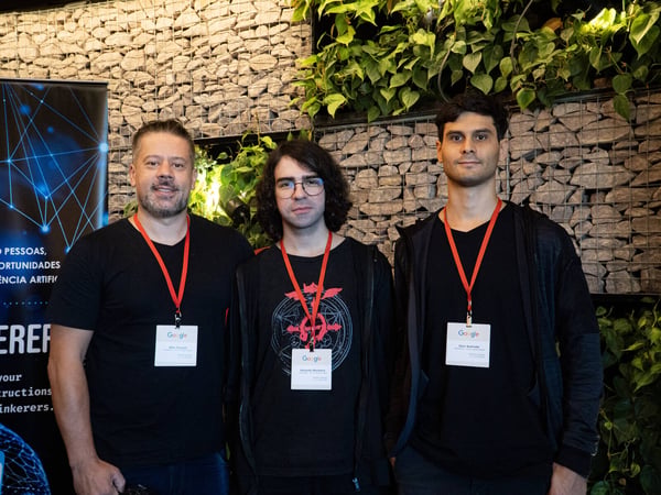 Three men wearing black shirts and Google ID badges stand side-by-side in front of a stone gabion wall and green plants.