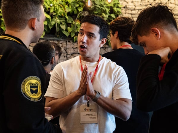 A group of young men are engaged in an animated discussion at a networking event, with the central figure speaking while holding his hands together.